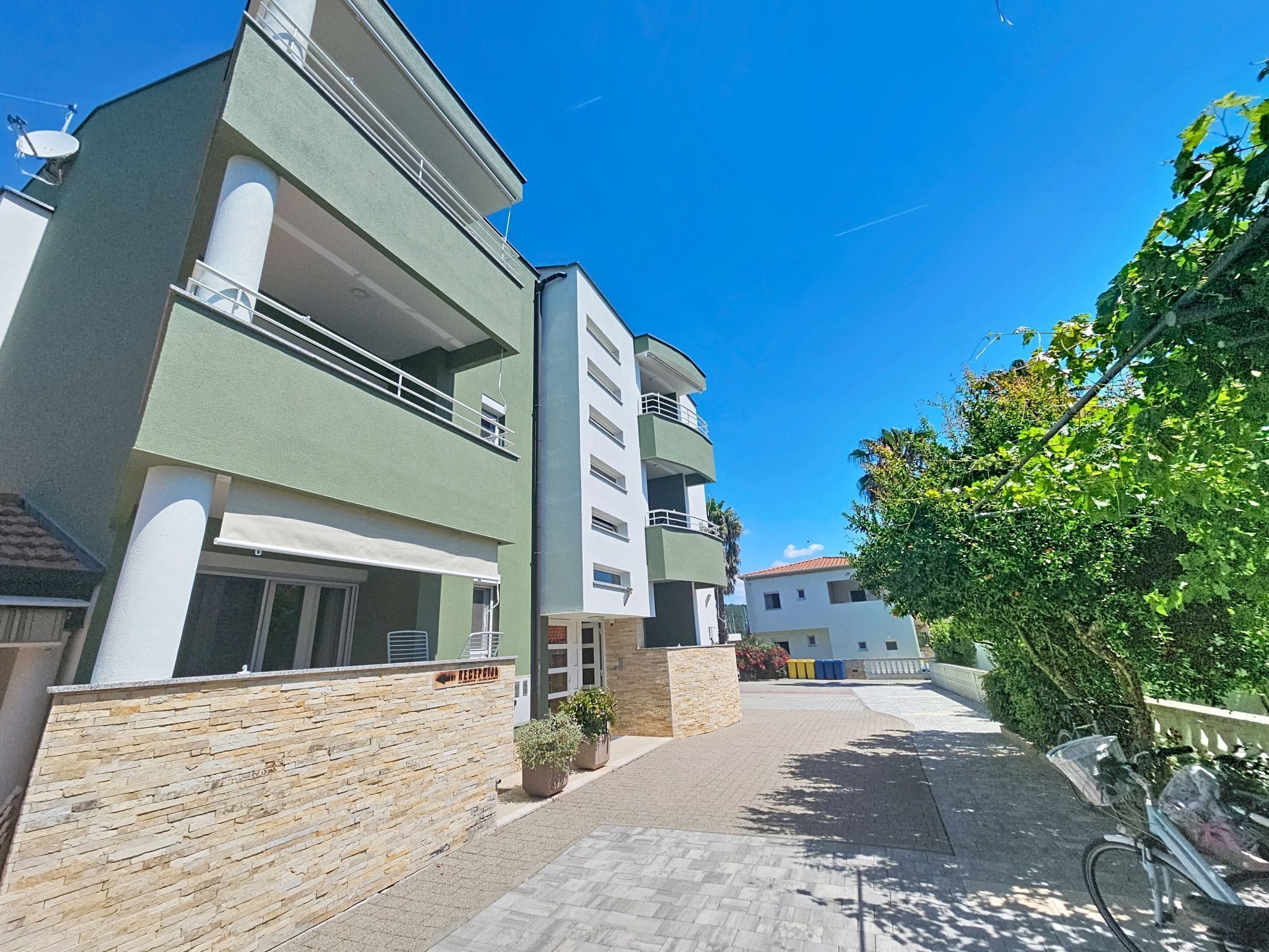 A multi-story residential building with balconies, a paved walkway, and a stone wall.