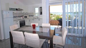 Una cocina moderna con electrodomésticos blancos, una zona de comedor con una mesa de vidrio y sillas blancas, y un balcón con vistas al mar.