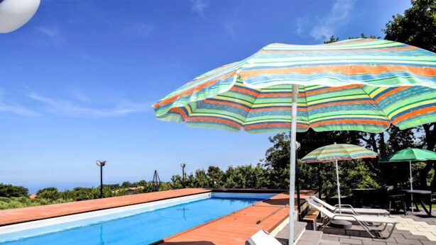 A swimming pool with lounge chairs and umbrellas, surrounded by greenery and a clear sky.