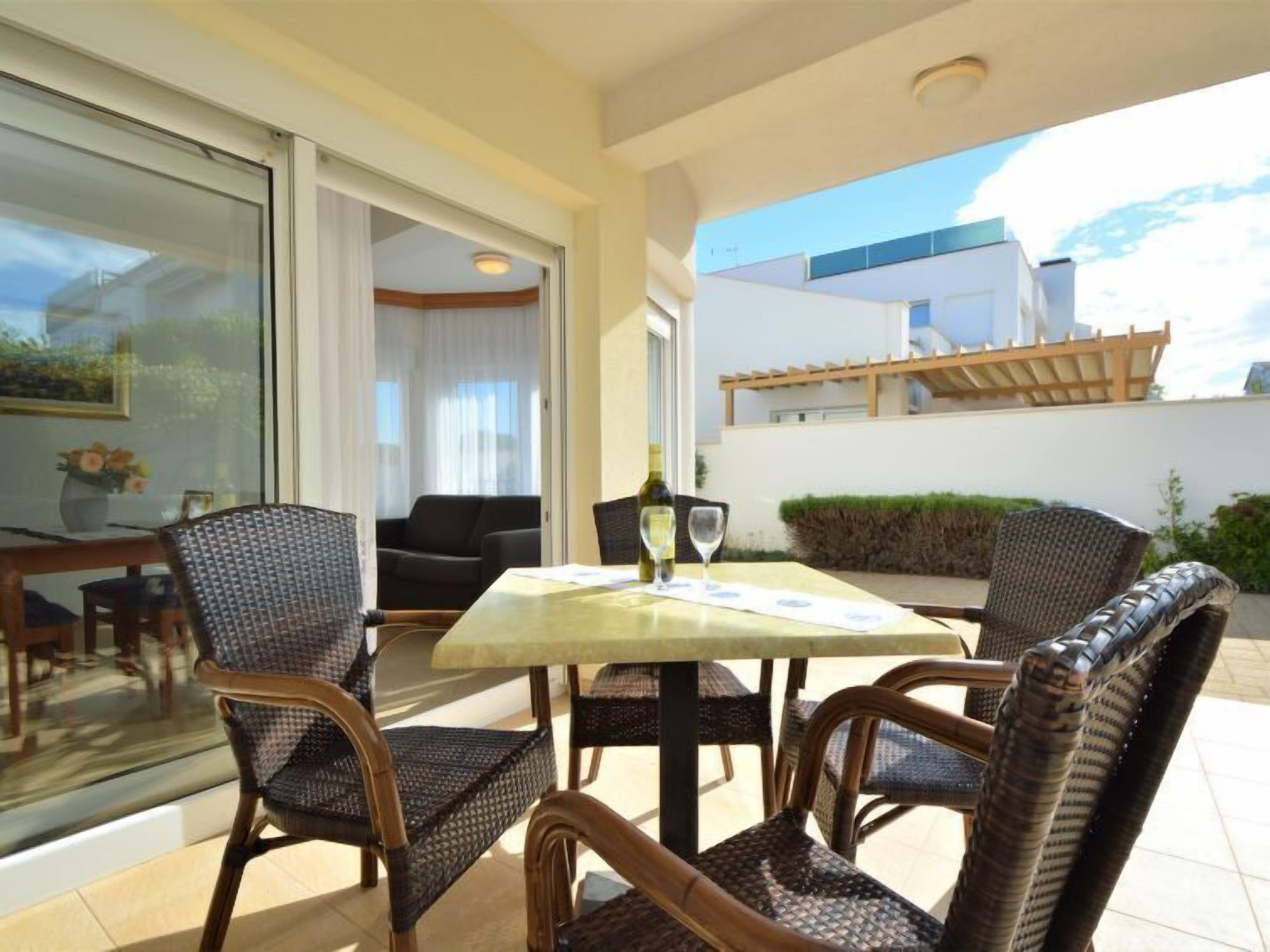 Un patio con sillas de mimbre y una mesa, una puerta de vidrio que da a una habitación, y vistas de un edificio.