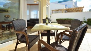 Eine Terrasse mit Korbstühlen und einem Tisch, eine Glastür, die zu einem Raum führt, und der Blick auf ein Gebäude.