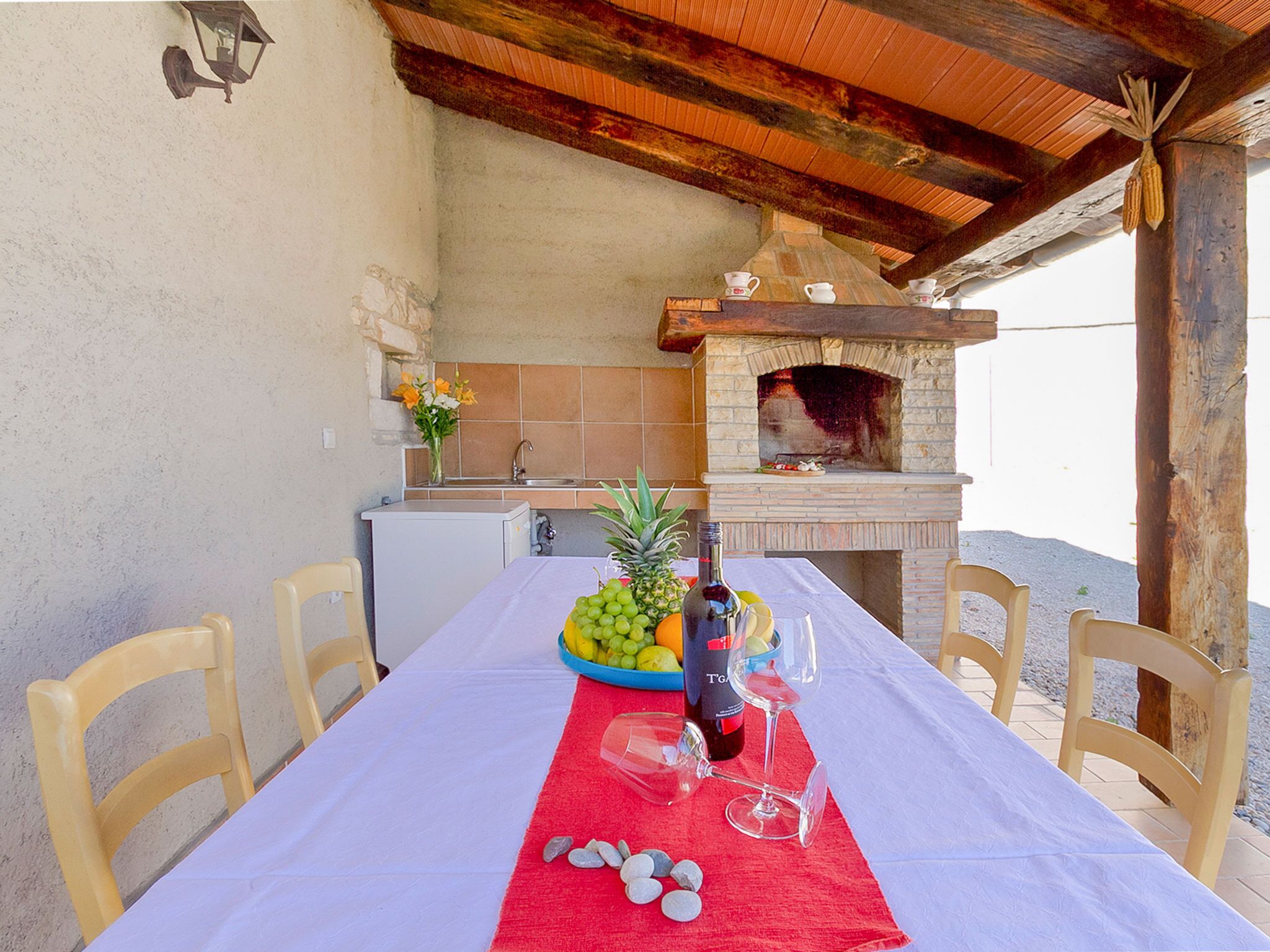 Un'area pranzo all'aperto con un tavolo imbandito per due, un forno a legna e un angolo cottura con frigorifero e lavello.