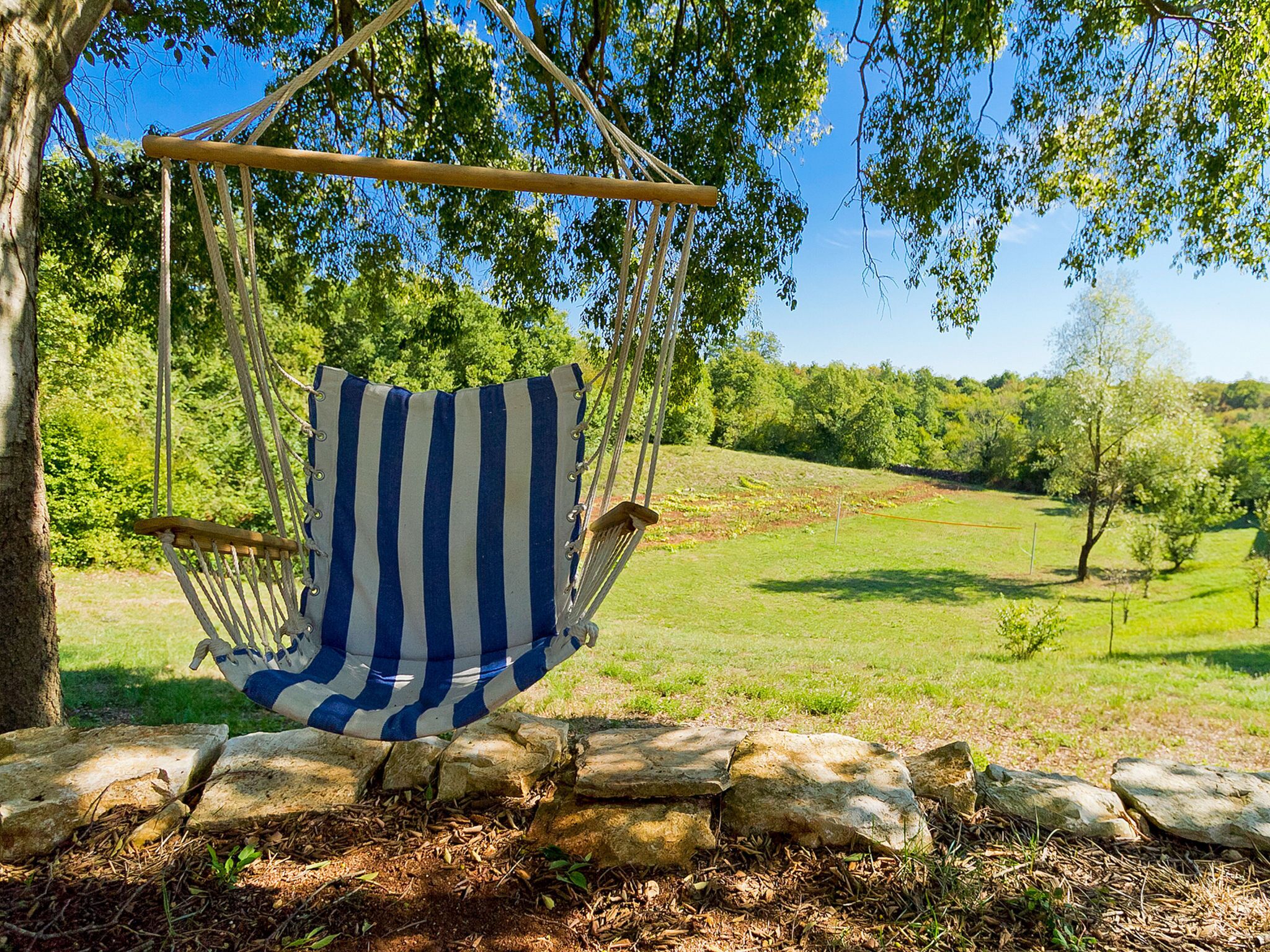 A hammock chair suspended from a tree in a grassy area with stone walls and trees in the background.