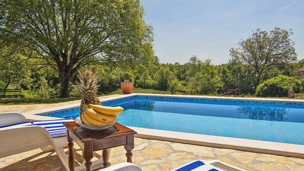 Un área junto a la piscina con sillones, una mesa con piña y plátanos, y un libro con gafas sobre una toalla.