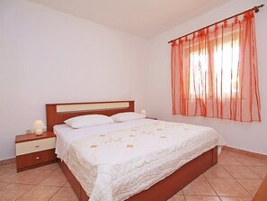 A bedroom with a wooden bed, white bedding, a nightstand with a lamp, and a window with sheer curtains. - Apartment Mladen by Interhome (Vodice)