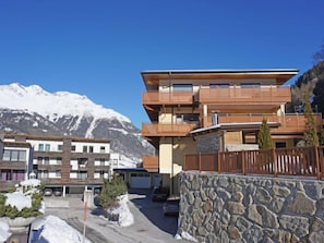 Un edificio de varios pisos con balcones, una pared de piedra y montañas al fondo.