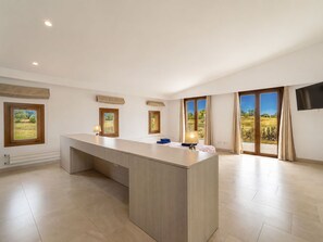 A spacious room with a large central island, a flat-screen TV mounted on the wall, and large windows with a view of greenery.