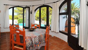 Uma sala de jantar com uma mesa redonda, seis cadeiras de madeira e amplas janelas em arco com cortinas.