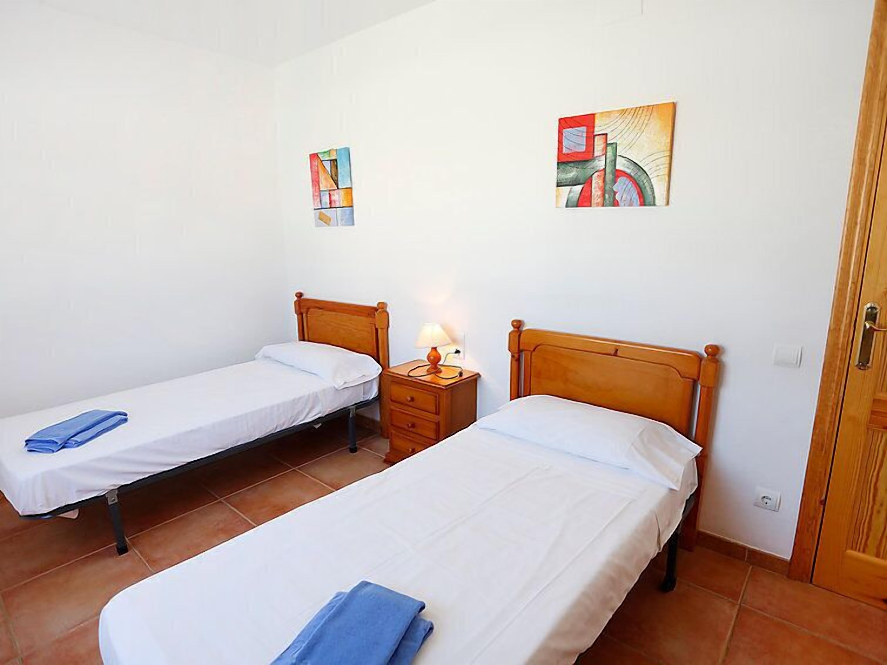 A room with two single beds, a nightstand with a lamp, and two abstract paintings on the wall.