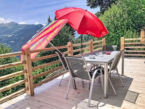 En træterrasse med et bord og stole, en rød parasol og udsigt til bjerge.