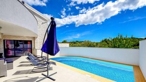 Un espace piscine aménagé sur le toit, avec des chaises longues et un parasol bleu, entouré d’un bâtiment blanc à la façade en pierre.