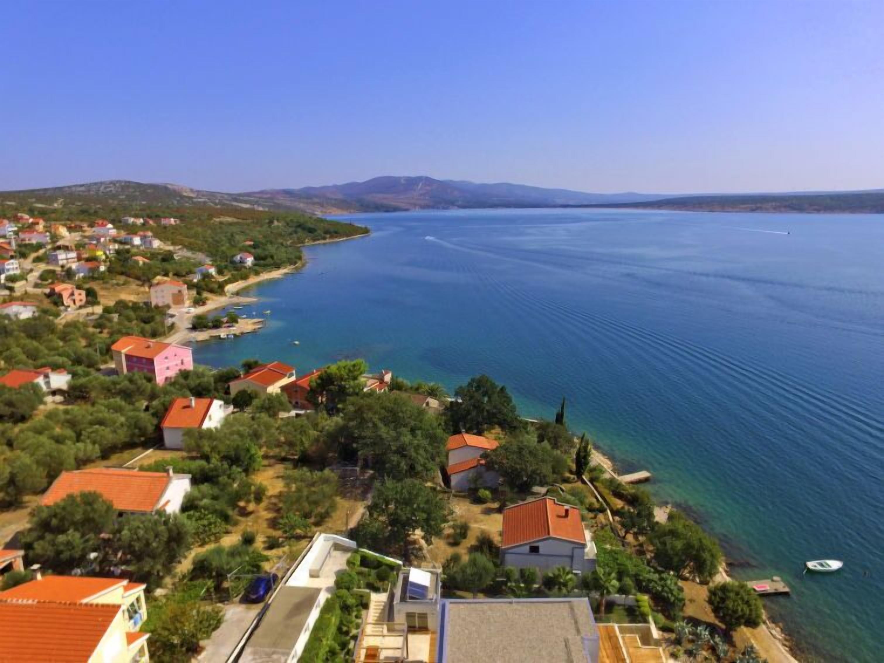 Un village côtier avec des maisons qui bordent le rivage, une mer d’un bleu limpide et des montagnes à l’horizon.