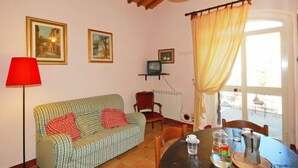 Uma sala de estar acolhedora com um sofá verde riscado, um abajur vermelho, uma mesa com uma cafeteira e um pote de vidro, e uma janela com cortinas.