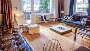 Ein modernes Wohnzimmer mit einer Ledersofa, einem weißen Sessel, einem grauen Sofa mit gemusterten Kissen, einem Couchtisch und einem Esstisch mit Stühlen. Große Fenster bieten Blick auf einen Balkon und die Außenanlage.