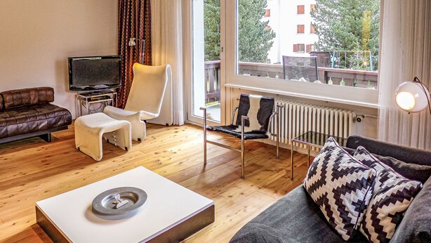 Ein modernes Wohnzimmer mit einer Couch, einem Sessel und einem Fernseher. Ein Fenster bietet Blick auf Gebäude und Bäume.