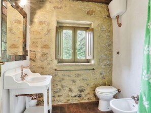 Ein rustikales Badezimmer mit Steinwand, Fenster, Waschbecken in Weiß, Toilette und Bidet.