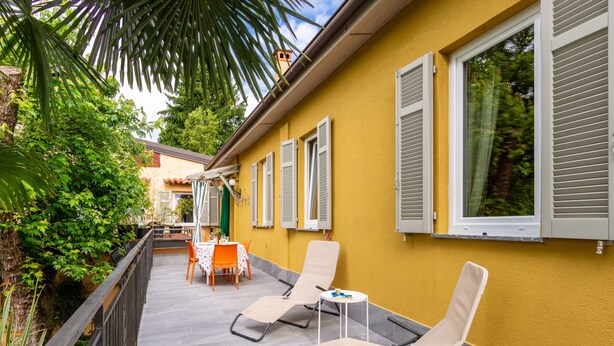 A yellow building with a balcony, outdoor seating, and a view of greenery.
