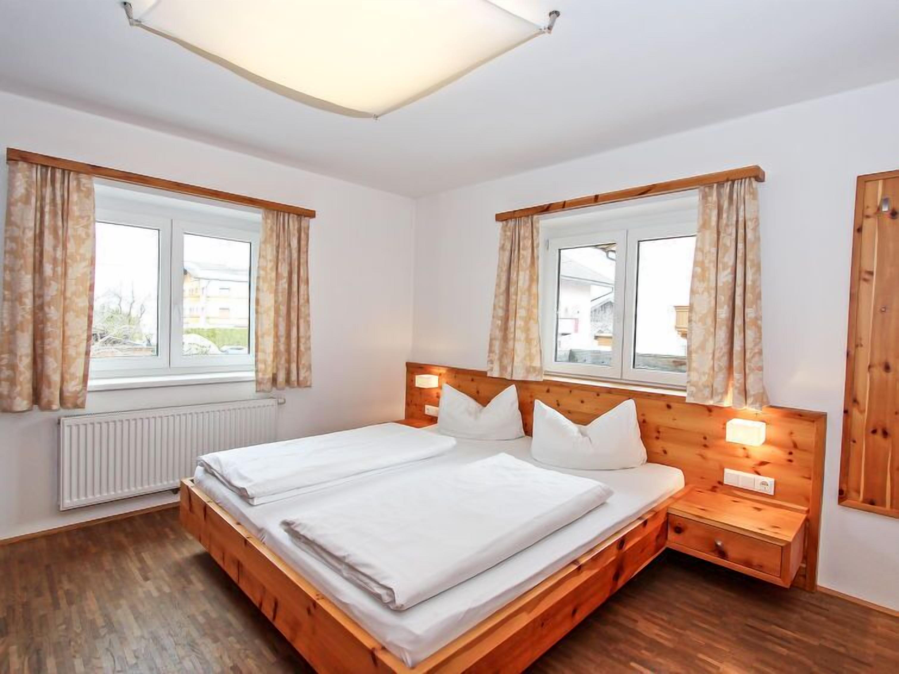 A double bed with wooden headboard and nightstands, two windows with curtains, and a radiator.