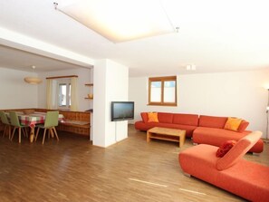 A modern living room with a wooden floor, a red sofa, a dining table with chairs, and a TV mounted on the wall.