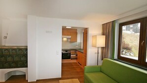 Een compacte leefruimte met een kitchenette, een groene bank en een raam met gordijnen.