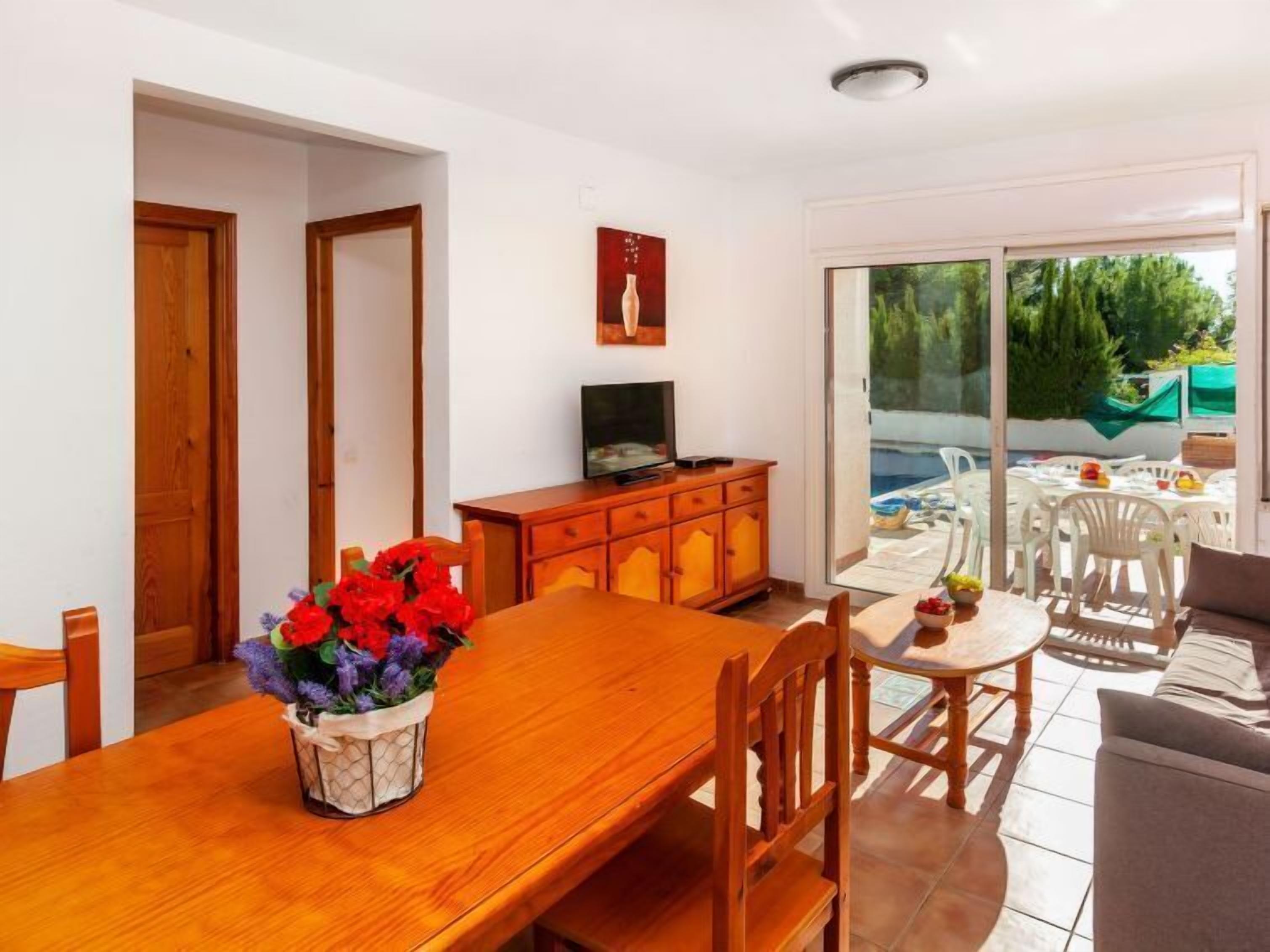 Un coin repas avec une table en bois, des chaises et un vase de fleurs. On y trouve également un téléviseur posé sur un meuble en bois, ainsi qu'une porte vitrée donnant sur un espace extérieur avec une piscine et des chaises.