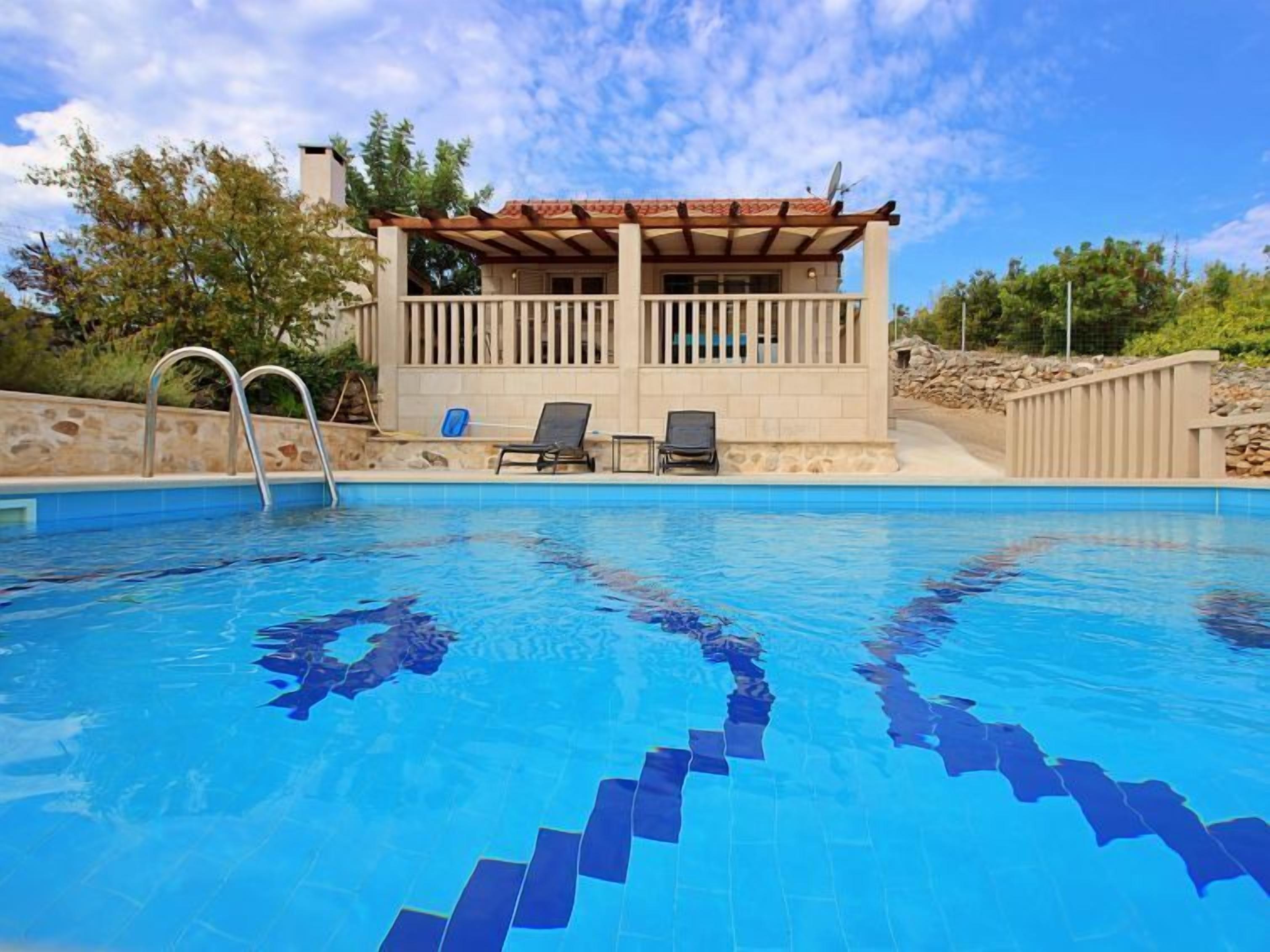 En swimmingpool med en klar, blå vandoverflade, et overdækket loungeområde med to liggestole og en stenvæg omkring poolen.