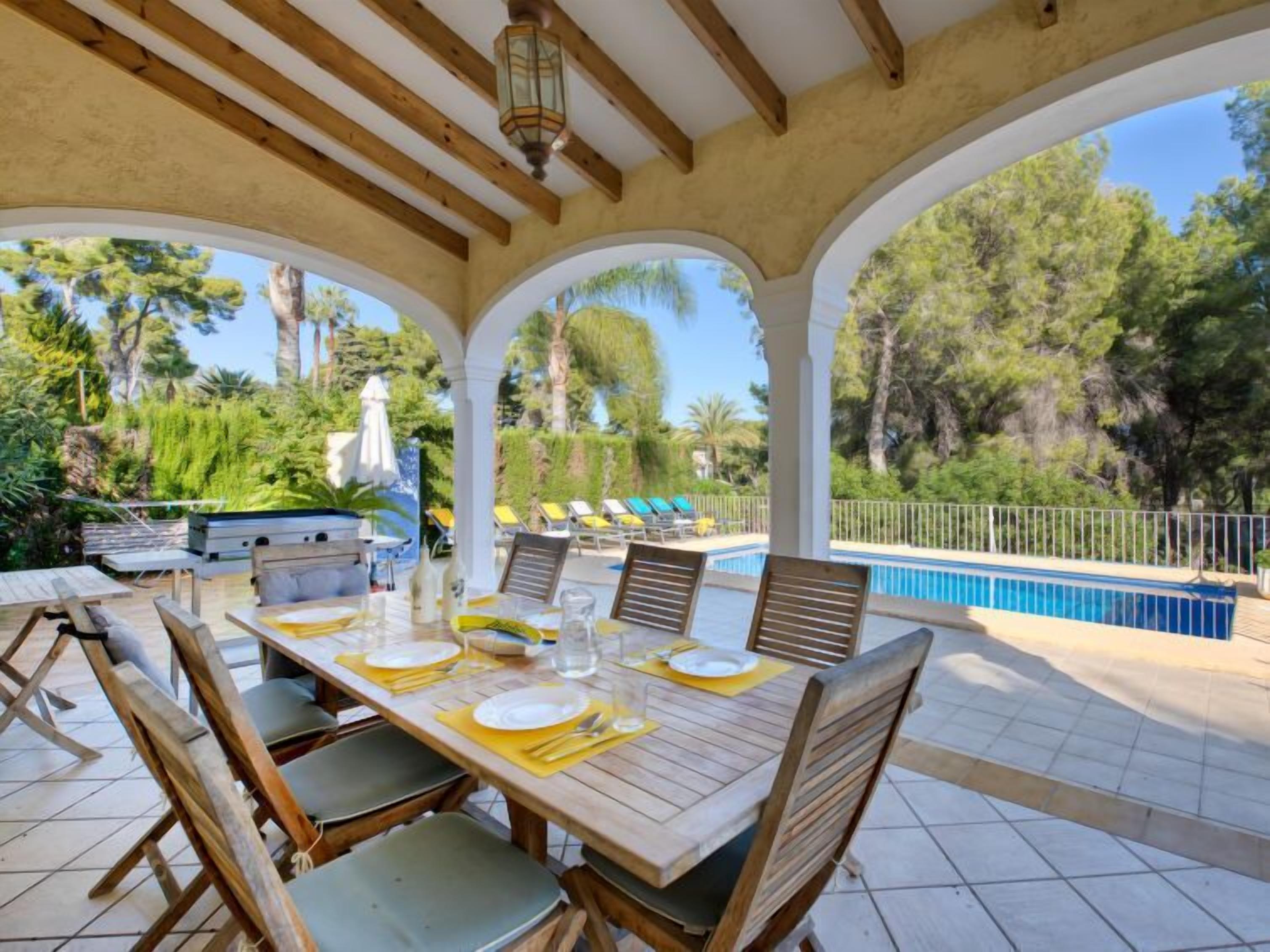 A covered outdoor dining area with a pool and lounge chairs in the background.