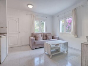 A spacious room with a light-colored tiled floor, a beige sofa with cushions, a wooden coffee table, and a window with curtains.