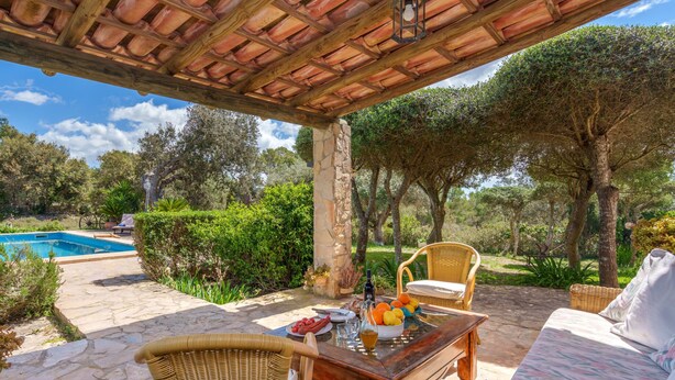 Eine Terrasse im Freien mit Steinboden, Korbmöbeln, einem Tisch mit Obst und Getränken sowie einem Pool im Hintergrund.