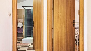 Un corridoio con pavimento in legno, cabina doccia in vetro e un letto con testiera bianca.