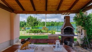 Una terrazza esterna con tavolo in legno apparecchiato per mangiare, pavimento in pietra e vista su un giardino sul retro con un barbecue.