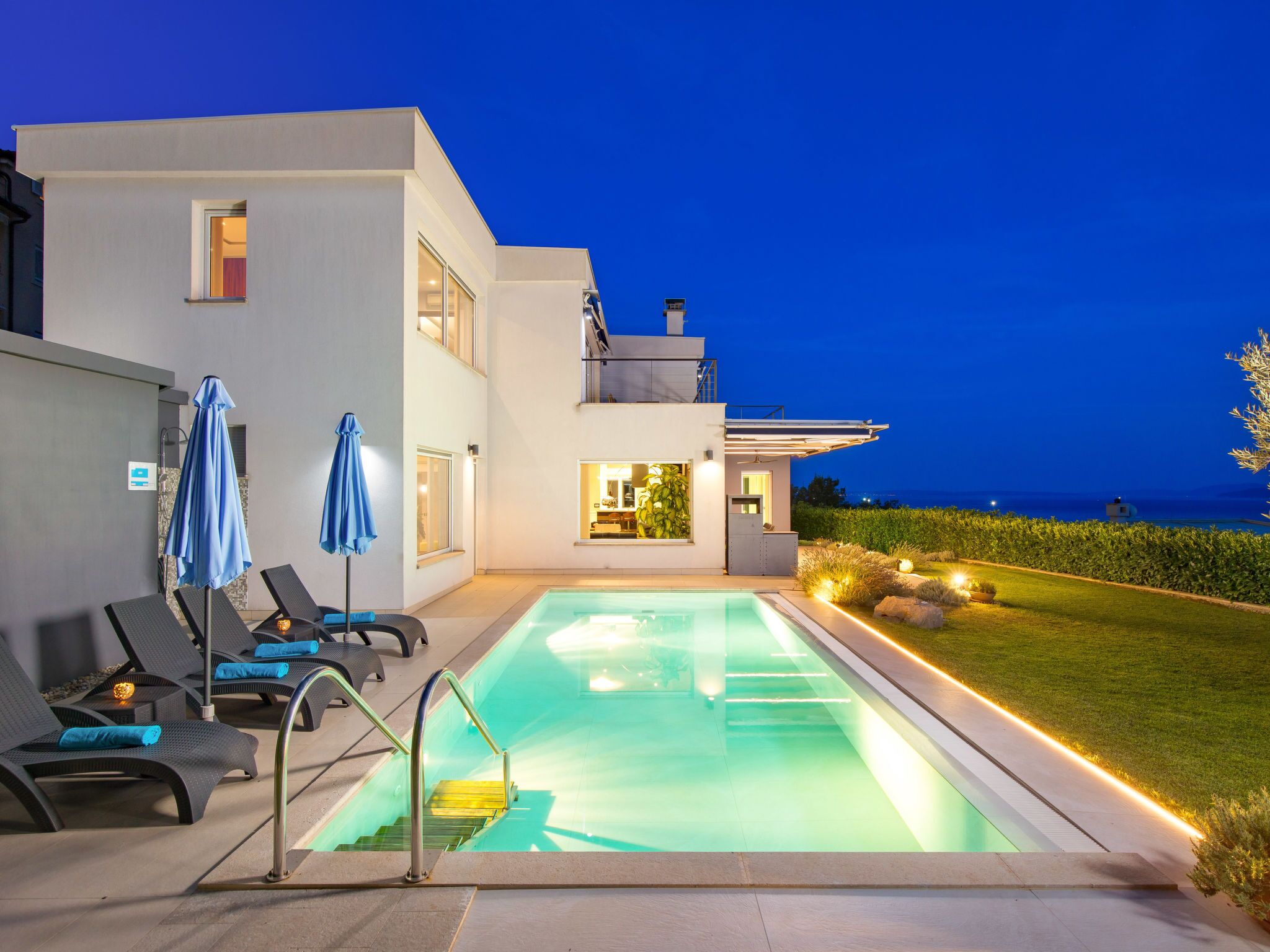A modern poolside area with lounge chairs and umbrellas, a clear pool, and a view of the sea at dusk.