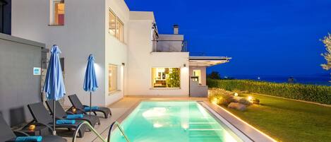 A modern poolside area with lounge chairs and umbrellas, a clear pool, and a view of the sea at dusk.