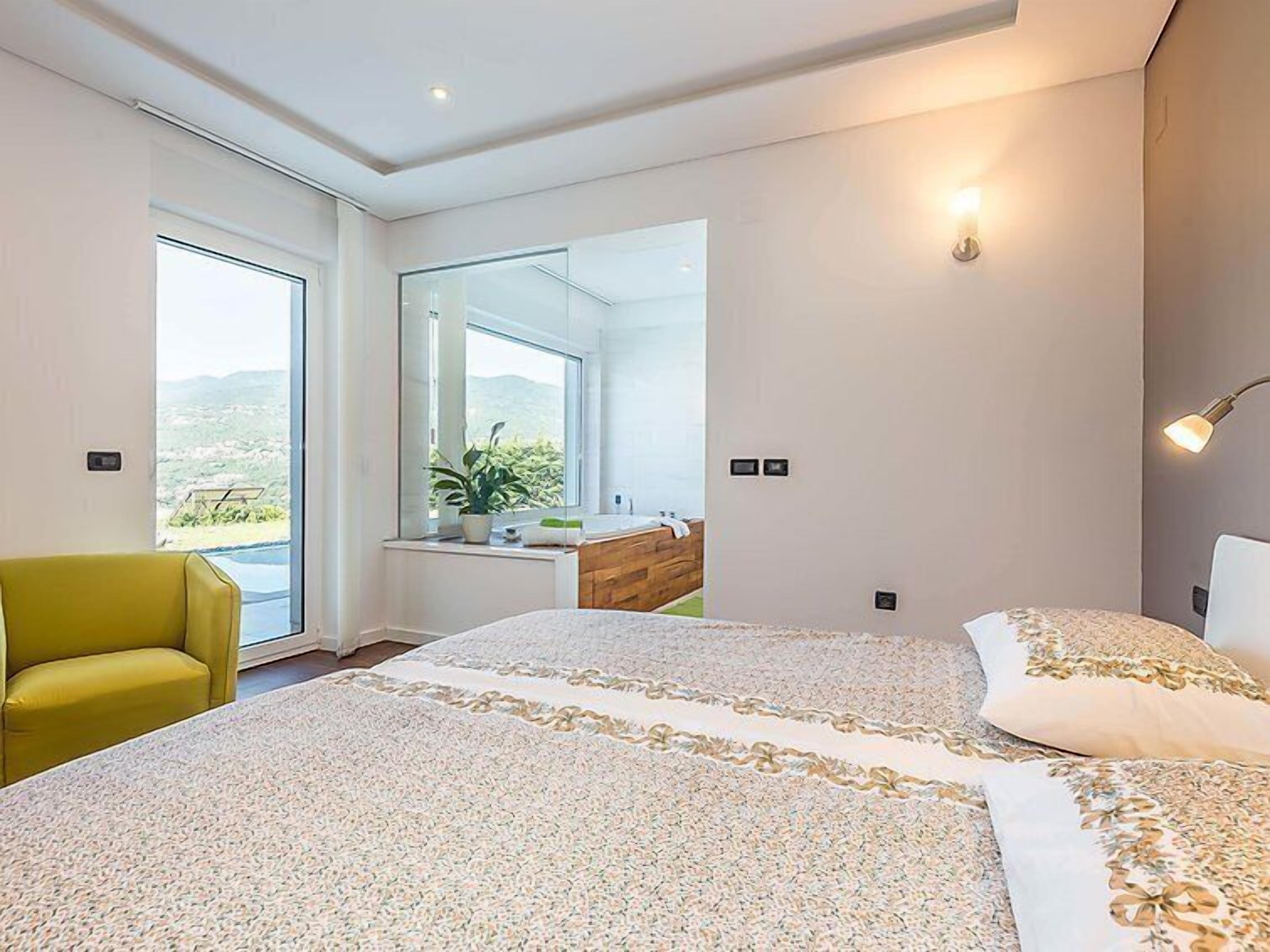 Een moderne slaapkamer met een groot bed, een groene fauteuil, een kaptafel met spiegel en uitzicht op het groen.