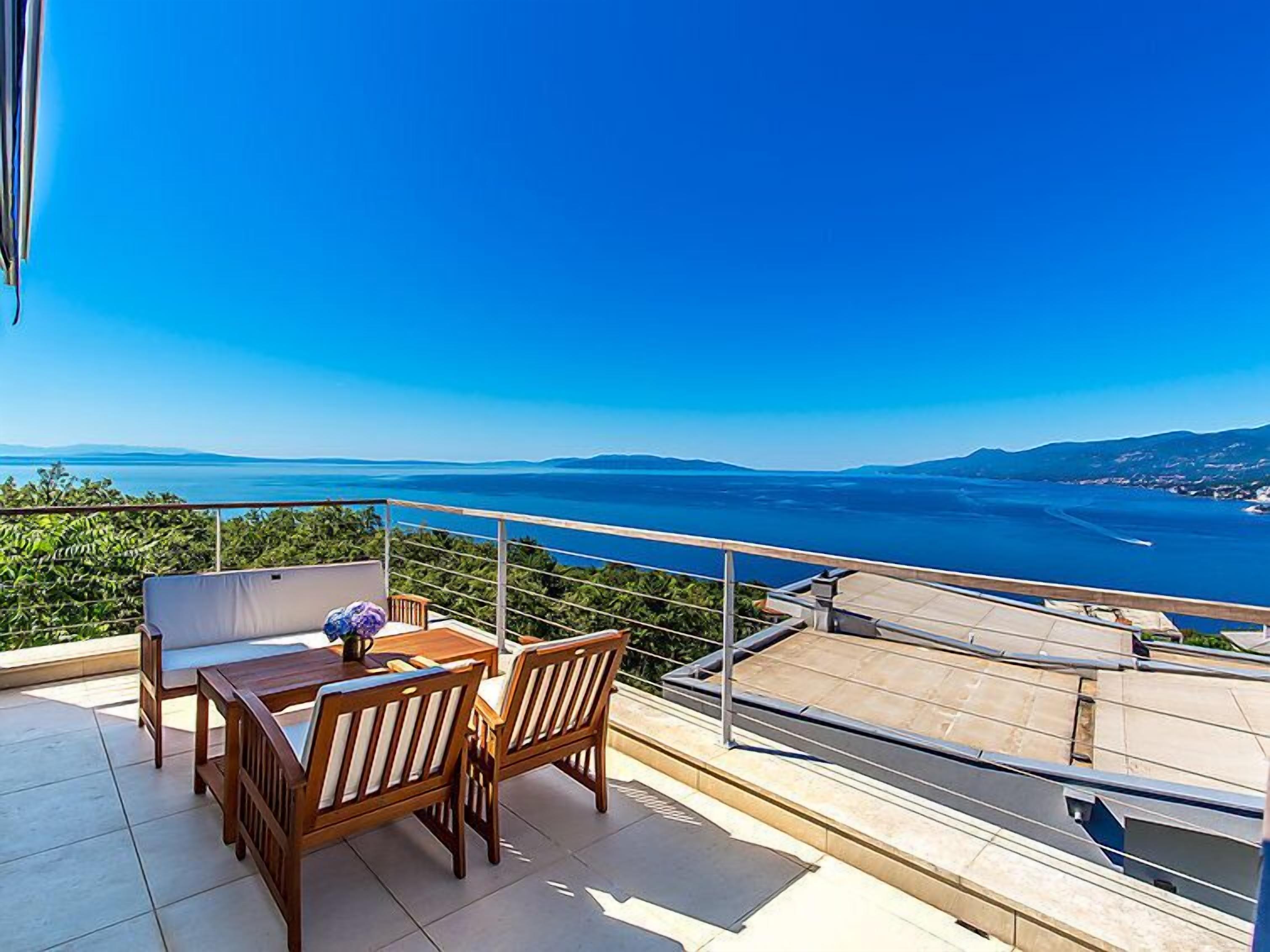 A balcony with a wooden table and chairs, overlooking a coastal view.