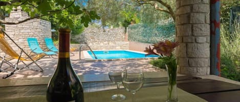 A bottle of wine, two glasses, and a vase with flowers on a table by a poolside.