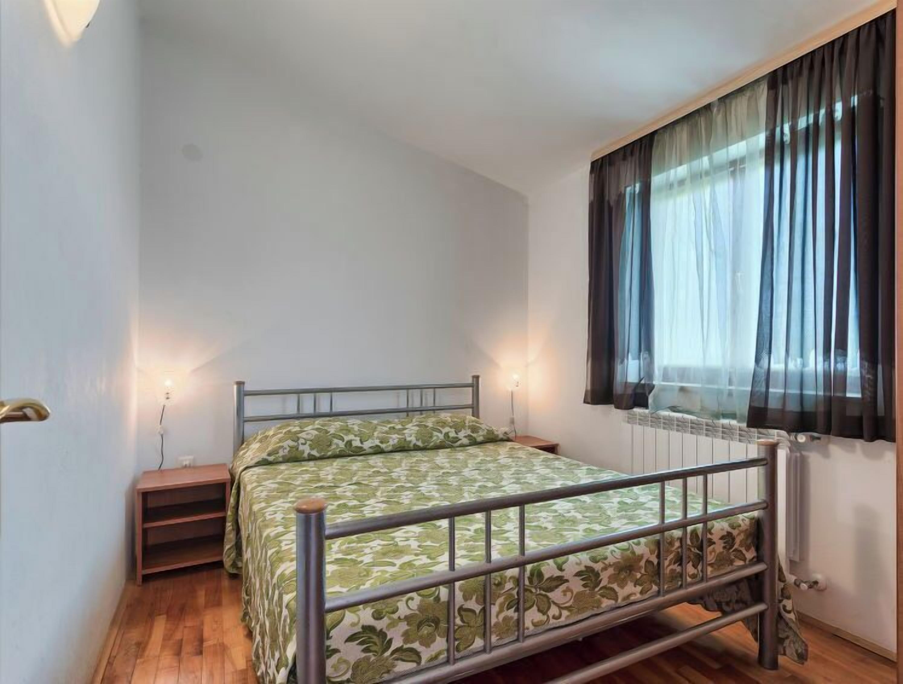 A small, single-bed room with a metal frame, a wooden floor, a nightstand, and a window with sheer curtains.