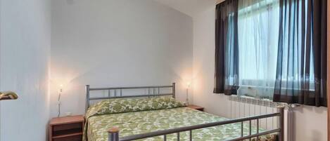 A small, single-bed room with a metal frame, a wooden floor, a nightstand, and a window with sheer curtains.