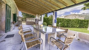 Een buitenterras met een marmeren tafel, rieten stoelen, een barbecue en uitzicht op een stenen gebouw en groen.