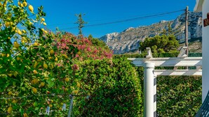Un jardin avec des citronniers et des fleurs roses, un bâtiment blanc et une montagne en arrière-plan.