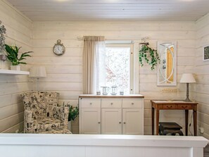 Une pièce avec un intérieur en bois peint en blanc, une armoire blanche avec des tiroirs, une horloge au mur, une fenêtre avec un rideau et une petite table avec une lampe.