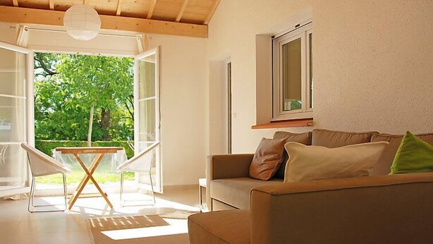 Una sala de estar acogedora con un sofá, una mesa y sillas, un ventanal con vista a follaje, e iluminación en el techo.