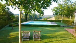 Una piscina con vistas a un paisaje montañoso, rodeada por un césped cuidado y dos sillas de madera.