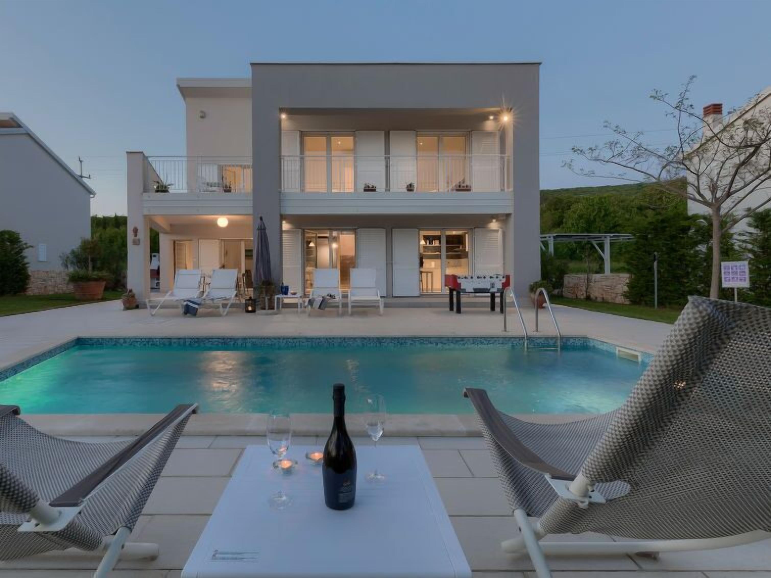 Ein zweistöckiges Haus mit Swimmingpool, Liegestühlen und einem Tisch mit Flasche und Gläsern.
