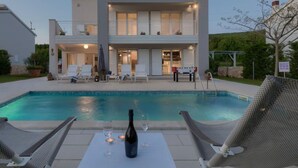 Ett tvåvåningshus med en swimmingpool, solstolar och ett bord med en flaska och glas.