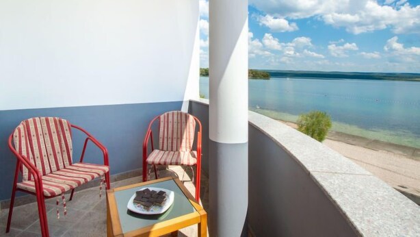 Un balcón con dos sillas a rayas, una mesa pequeña con un plato de comida, y una vista de un lago tranquilo bajo un cielo despejado.