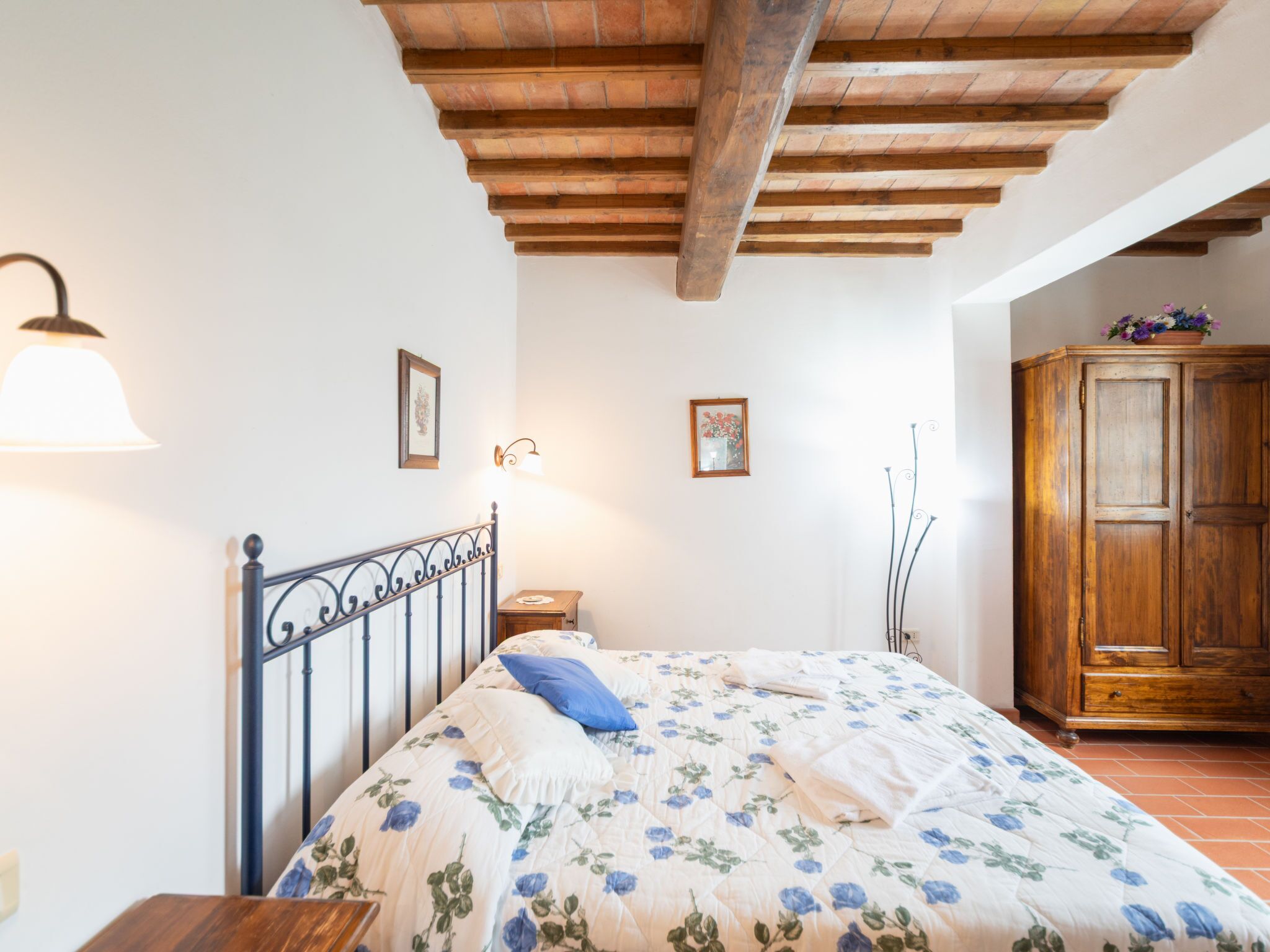 A bedroom with a wooden ceiling, a bed with floral bedding, a nightstand, a lamp, a wooden wardrobe, and a wall-mounted picture.