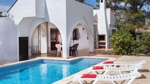A poolside area with a white building featuring arched doorways and a small patio with a table and chairs. - Villa Zozo by Interhome (L'Ametlla de Mar)