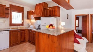 A kitchen with wooden cabinets, a granite countertop, a refrigerator, and a bowl of fruit. - Villa Zozo by Interhome (L'Ametlla de Mar)