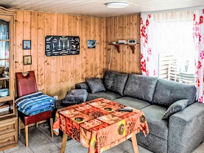 Una sala de estar acogedora con una pared de madera, un sofá, un sillón, una mesa con mantel floral y una estantería.